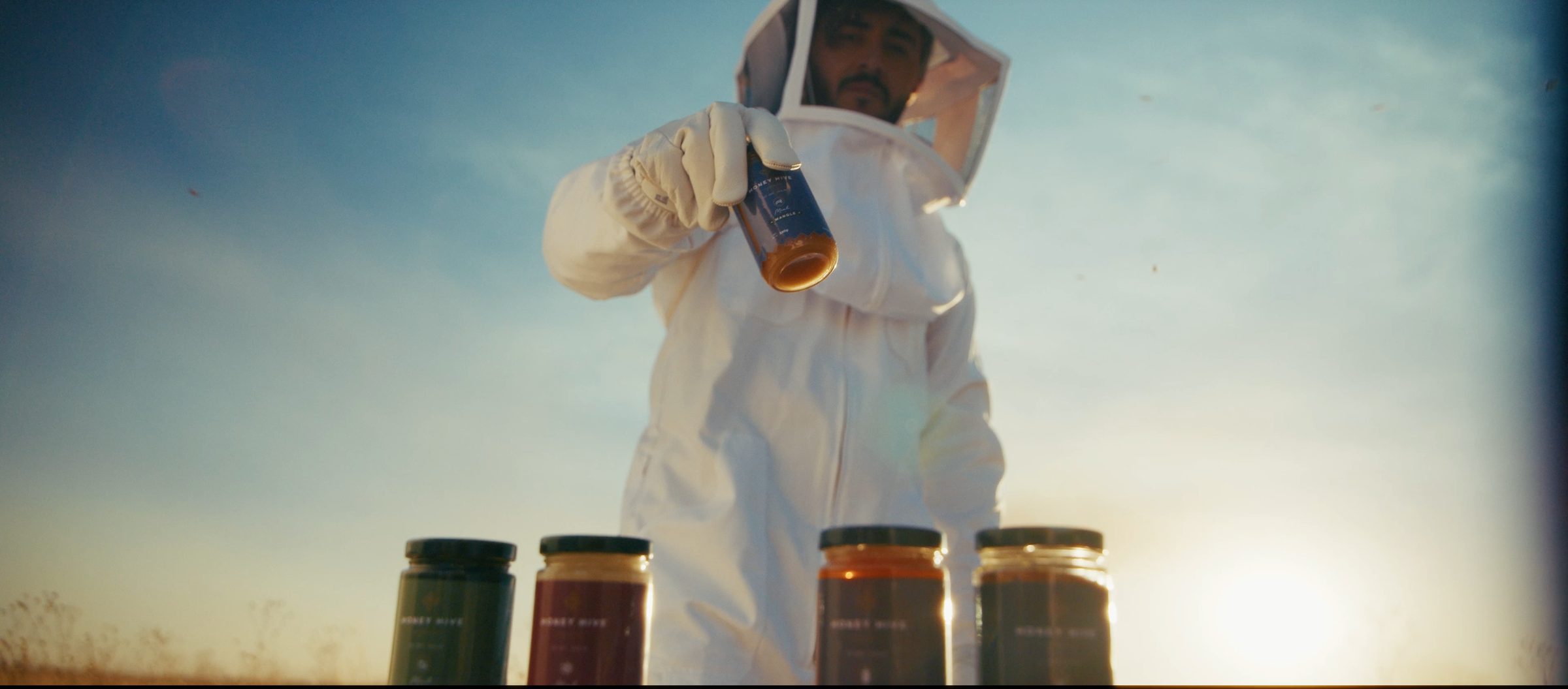 Apicultor con traje blanco sirve miel junto a cuatro frascos al atardecer